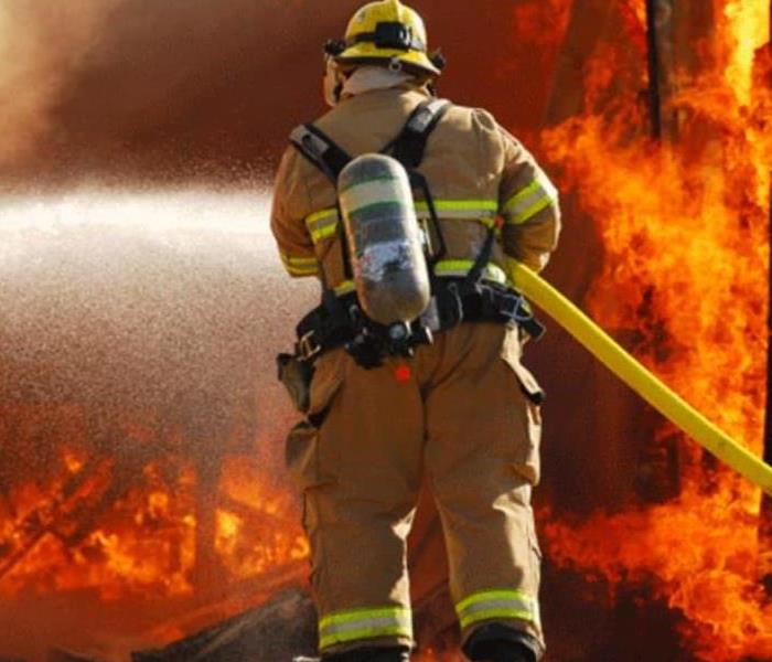 This firefighter is dressed in uniform with a fire infornt of him, water hose in hads and spraying the fire.